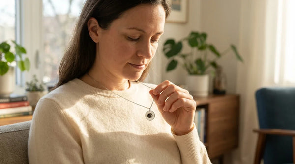 A woman seated near a sunlit window looking down softly at a delicate Brother & Sisters Circle Photo Necklace pendant resting against her cream sweater, golden hour light