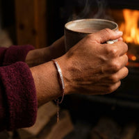 Thumbnail for Crystal Bar Bracelet or rose porte au poignet, mains autour d'une tasse devant un feu