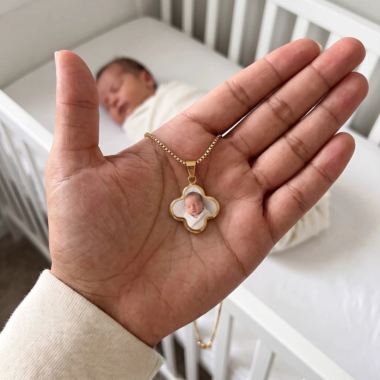 The gold clover photo pendant in an open palm, real small size, a newborn sleeping in the crib behind | SKU: TPH-CLVN-GOLD